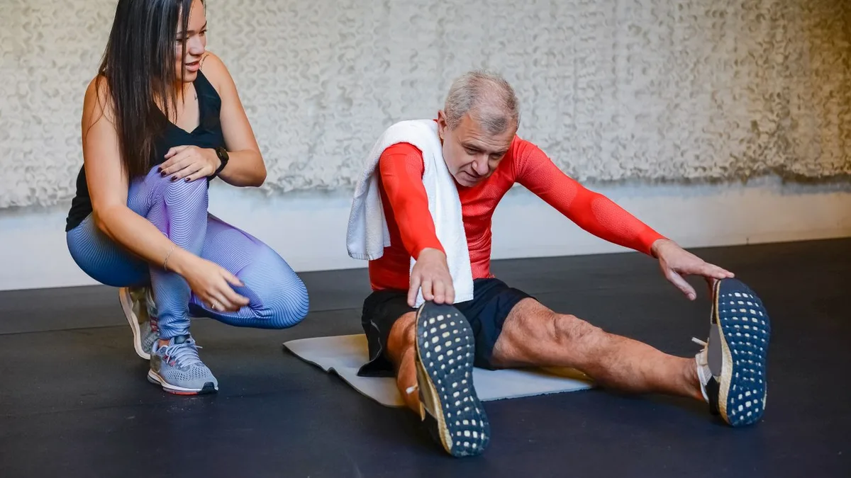 An older man stretching on a mat with a trainer's assistance.