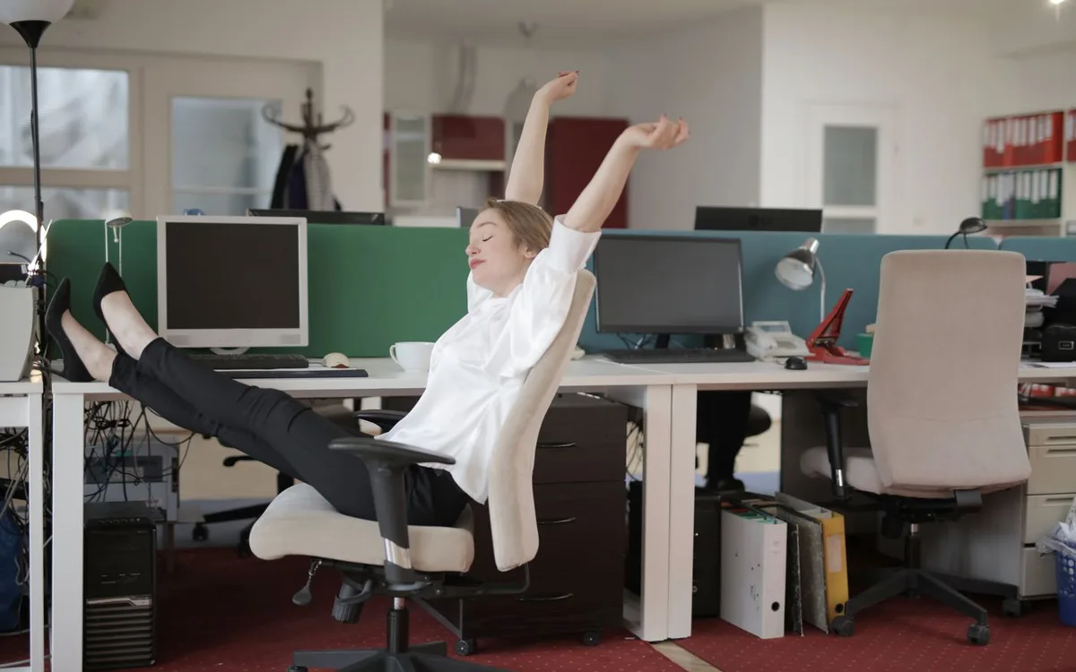 An office worker raises her arms while seated at a desk with computer monitors.